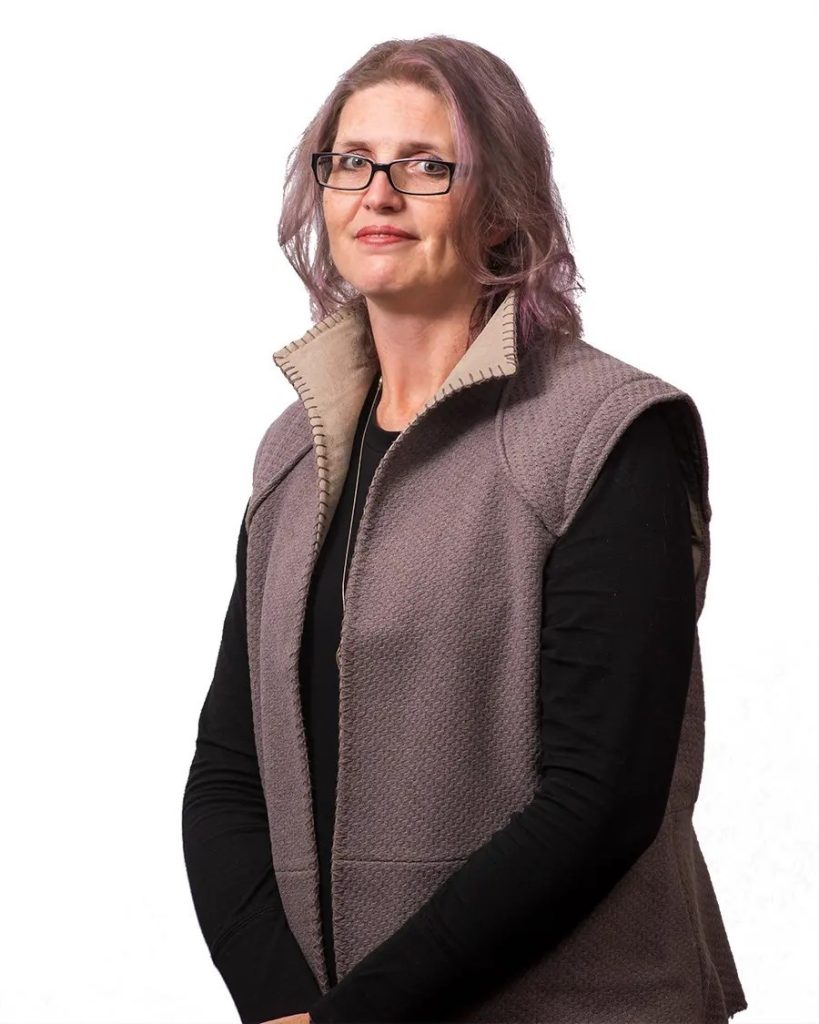 A photo of C. Aaron Sharrock on a white background. Sharrock is a white woman with shoulderlength ash blonde hair, wearing glasses and a black longsleeved shirt under a brown vest.