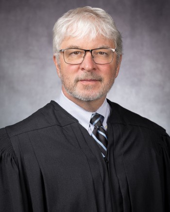 A headshot of Justice Michael P. Donley on a gray background. Justice Donley is a white man with white hair, mustache, and beard, wearing glasses, a black and blue striped tie, and white shirt under a black judicial robe.