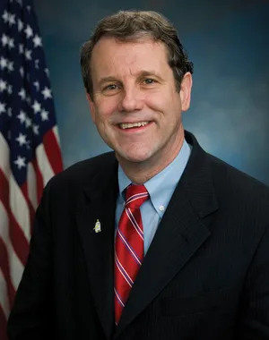 A headshot of US Senator Sherrod Brown in front of an American flag. Senator Brown is a white man with dark gray hair, wearing a red and white striped tie, blue colored shirt, and a black suit jacket.