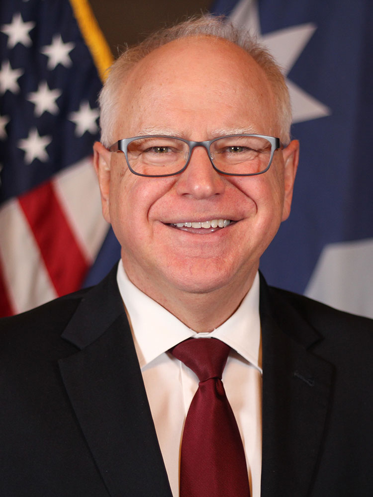 A headshot of Governor Tim Walz in front of an American flag. Governor Walz is a white man with white hair, wearing glasses, a red tie, and a white collared shirt under a black suit jacket.