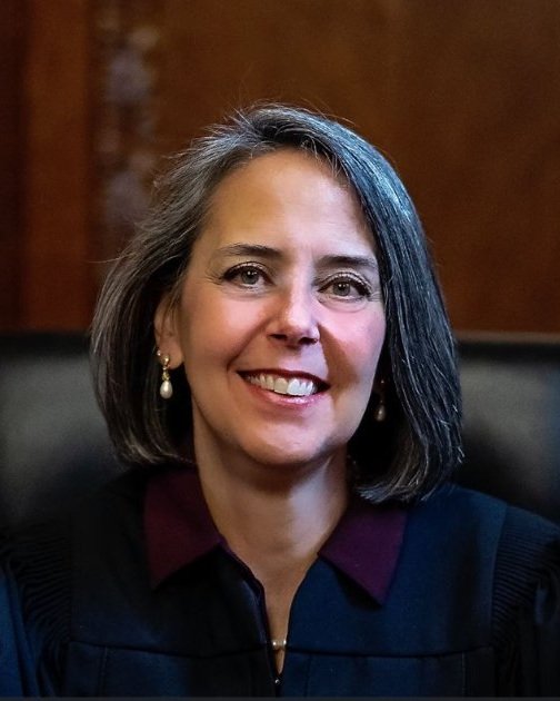 A headshot of judge Lisa Forbes sitting in a black chair in front of a brown wall. Judge Forbes is a white woman with shoulder-length salt-and-pepper hair, wearing pearl jewelry and a burgundy collared shirt under a black judicial robe.