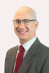 A headshot of Craig Swartz on a white background. Swartz is a white man with white hair, wearing glasses, a red tie, and a white collared shirt under a gray suit jacket.