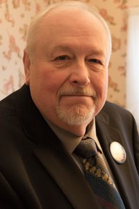 A headshot of Keith Mundy in front of a floral wallpaper background. Mundy is a white man with white hair, mustache, and goatee, wearing a blue tie and gray shirt under a black suit jacket.