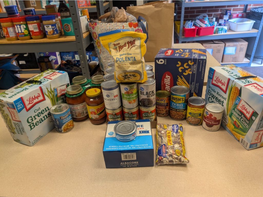 A table full of canned goods, dried beans, dry pasta, pasta sauces, and a bag of polenta is in front of shelves full of other nonperishable food items.
