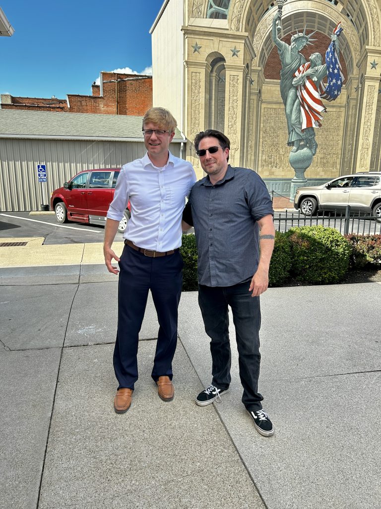 Bryan Hambley and Chris Volpe stand closely with an arm behind each other's back. The background shows a mural of the Statue of Liberty holding a person draped in the American flag.