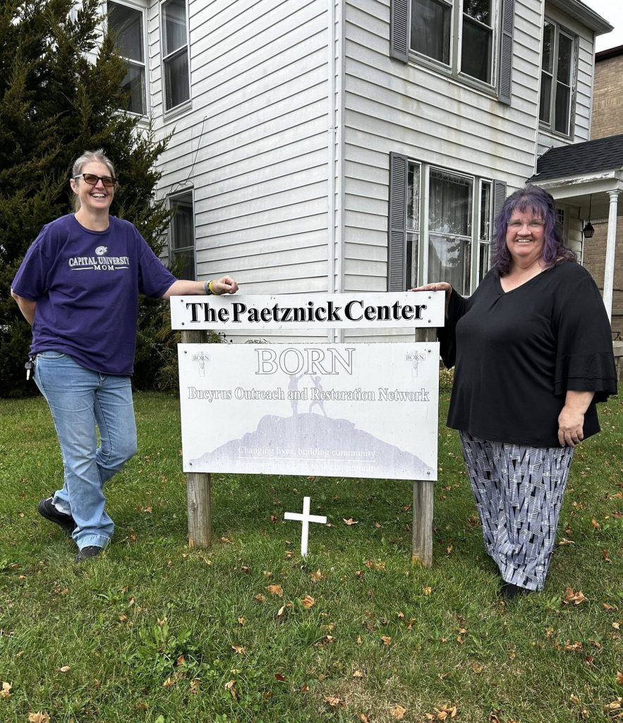 Two women stand in front of a white house with black shutters. Between them, is a sign that reads, “BORN; Bucyrus Outreach and Restoration Network; Changing Lives, Building Community, Representing Christ to the Community.” A cross is posted in the ground under the sign.