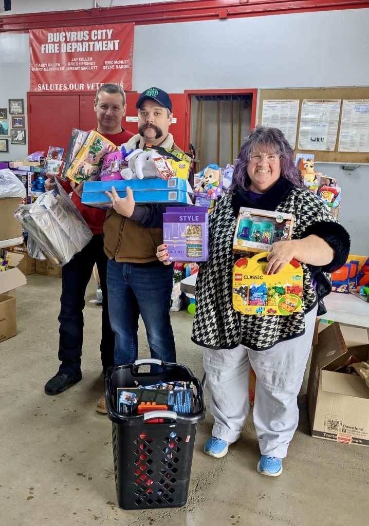 Two men and a woman with arms full of toys stand in a row. Boxes and baskets of toys surround them. A red banner behind them reads: “Bucyrus City Fire Department."