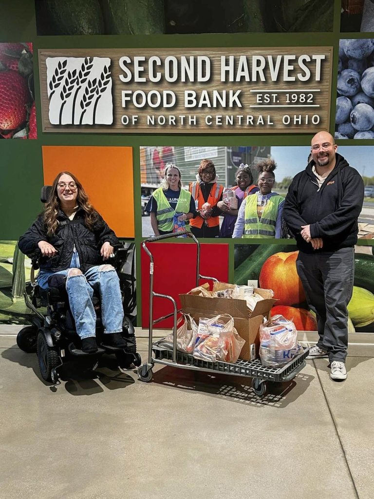 A woman in a wheelchair and a man are in front of a mural, which contains photos of fresh fruits and vegetables, volunteers, and a sign that says, “Second Harvest Food Bank of North Central Ohio. Est. 1982." A box and bags of shelf-stable food sit on a cart between them.