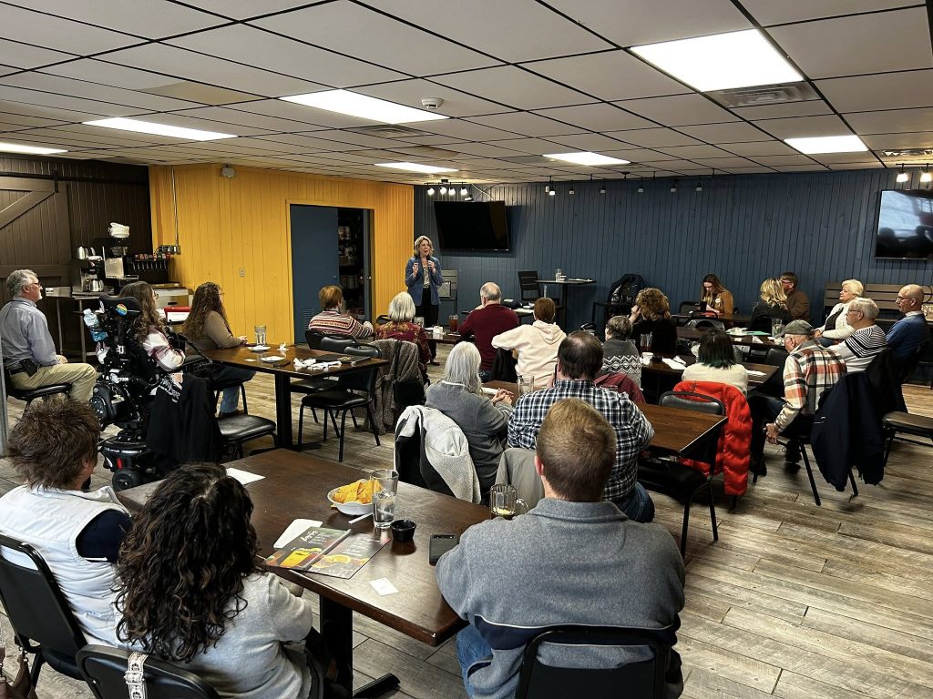 A community gathered at tables within a room with yellow and blue walls. Alison Russo stands in front of the crowd.
