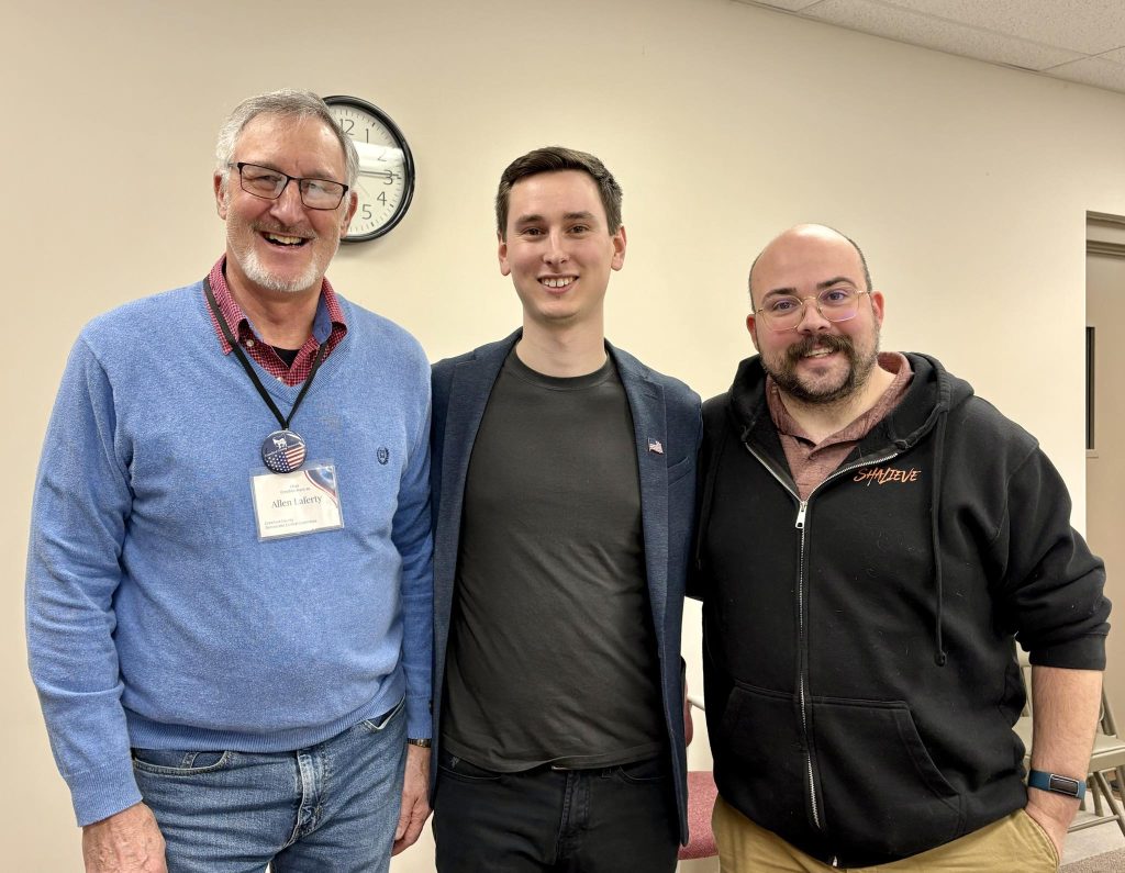 Allen Laferty, Daniel Burket, and Seth Bennett are smiling and standing in a row. The background is a beige wall in a casual meeting room with foldable chairs visible behind the men. 