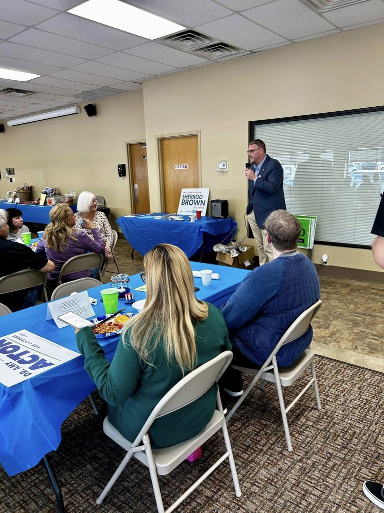 A man speaks into a microphone at the front of a room with seated attendees. Tables are covered in blue cloths, creating a casual and engaging atmosphere.