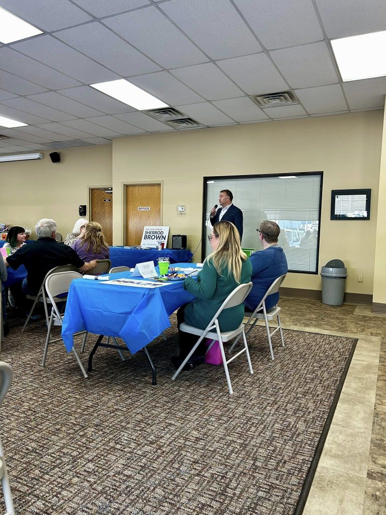 A man speaks into a microphone at a community event with people seated at tables covered in blue tablecloths. The atmosphere is engaged and casual.