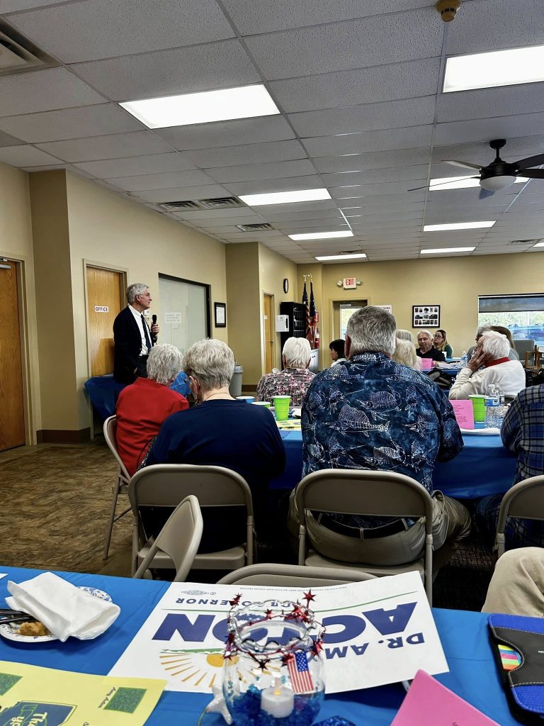 A man in a suit speaks at a community gathering in a meeting room. Attendees sit at tables with colorful papers, creating a warm and engaged atmosphere.
