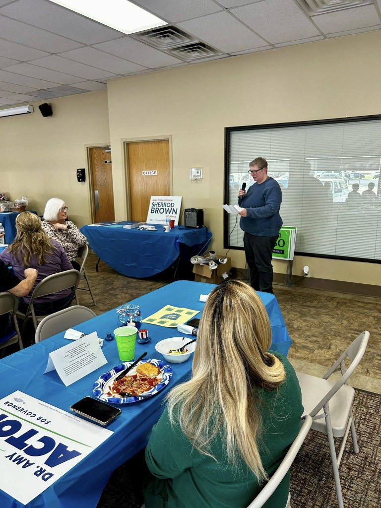 A person speaks into a microphone in a room with blue table covers and campaign signs. Seated individuals listen attentively. The atmosphere is focused.