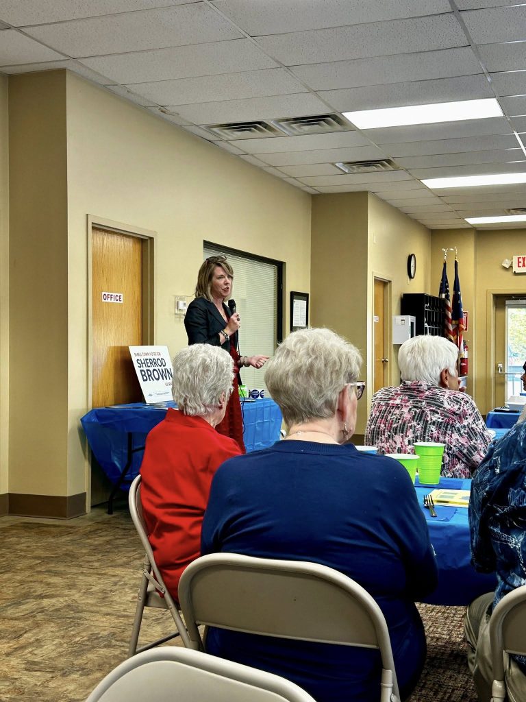 A woman speaks into a microphone in a community room with a sign for Sherrod Brown. Seated audience members listen attentively.