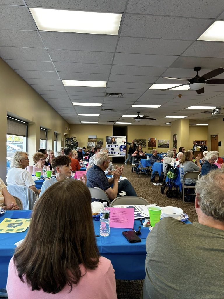 A community gathering in a bright room with people seated at tables covered with blue cloths. The mood is attentive.